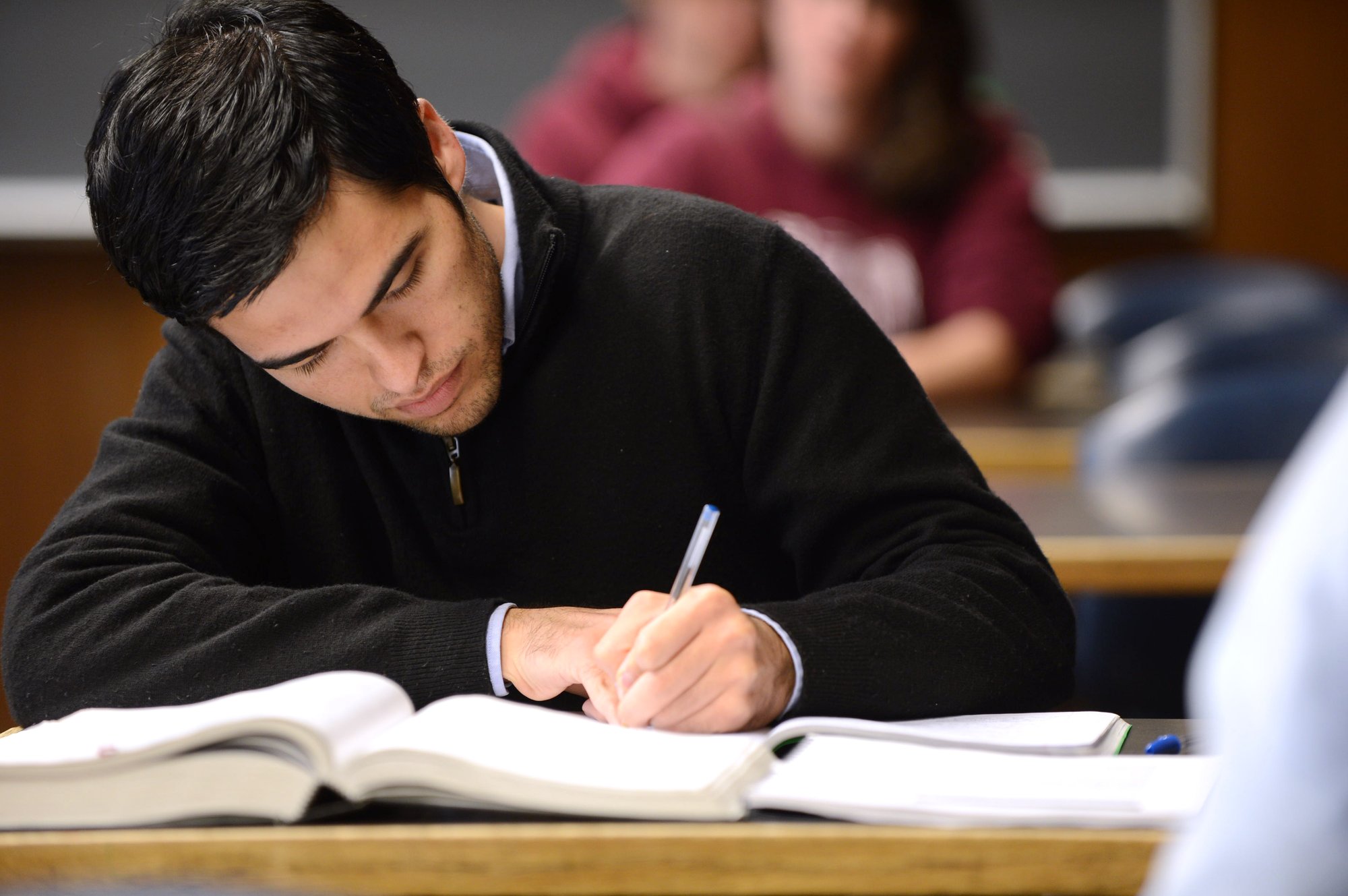 A man is writing in a notebook while sitting at a desk with an open book in a classroom setting.